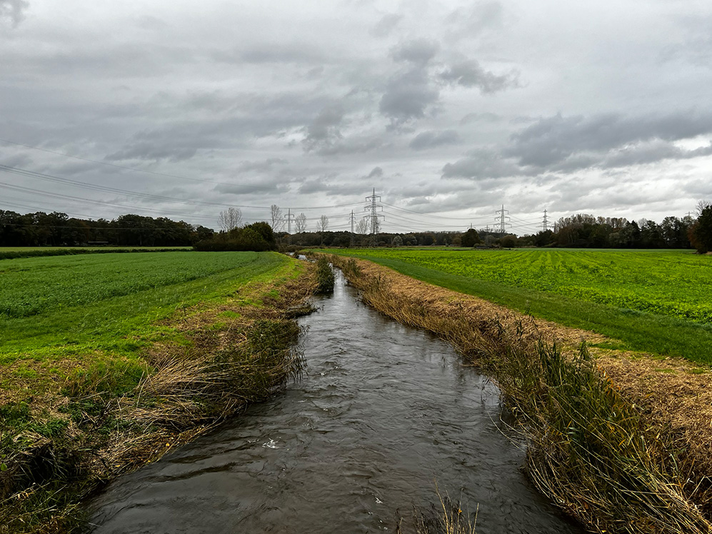 Zu sehen ist ein Fluss, der durch Felder fließt vor einem stürmischen Himmel. 